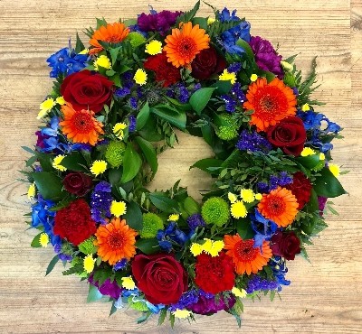 Elegant funeral wreath with vibrant flowers in orange, red, blue, purple, green, and yellow, accented with lush foliage.