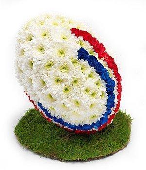 3D rugby ball funeral tribute with white chrysanthemum base, red and blue spray paint detailing, turf-effect base, and optional team colours or logo.