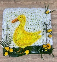 Duck-shaped funeral tribute made with white chrysanthemum, detailed with spray-painted shading and finished with foliage and yellow floral accents on a 2ft x 2ft base.