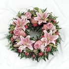 Soft pink funeral wreath with lilies, roses, chrysanthemums, and carnations, accented with foliage and grass loops.